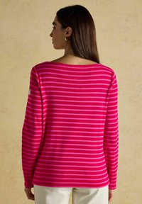 Vaaleanpunainen ja magentanauhoituksilla varustettu pitkähihaisella puserolla, jossa on leveä pääntie. Kangas näyttää pehmeältä ja hieman tekstuuriselta, ja hihassa on pieni logo.