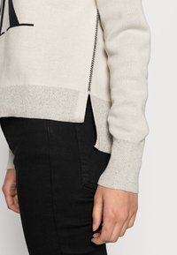 Gros plan sur une personne portant un pull crème fendu sur les côtés et un jean noir, montrant sa main gauche détendue le long du corps.