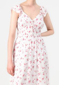 Nainen, joka käyttää valkoista mekkoa, jossa on vaaleanpunaisia kukkakuoseja. Mekossa on lyhyet liehuvat hihat, syvä V-pääntie sekä kerätty vyötärö, jossa on nauhakoristelu.