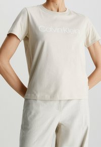 Beige bomull t-shirt med rund halsringning och korta ärmar, med vit "Calvin Klein" text över bröstet. Slät textur.