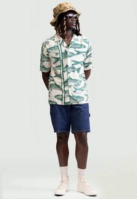 Camisa de manga curta com estampado de peixe em verde sobre um fundo creme, combinada com calções de ganga escuros e sapatilhas creme. Chapéu de excursão.