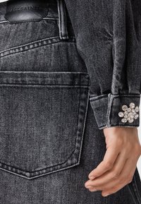 Chaqueta de mezclilla en color carbón con un acabado texturizado, que presenta un acento de flor de cristal en el puño y una etiqueta de cuero en la parte posterior.