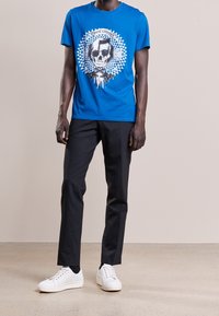 Blå bomullst-shirt med grafik av ett skellet och geometriskt mönster, kombinerad med svarta byxor och vita sneakers. Korta ärmar, rundad halsdesign.