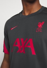Szary sportowy T-shirt z krótkim rękawem. Na piersi znajduje się duży czerwony napis "AXA", czerwone logo Nike oraz czerwista odznaka Liverpool FC.