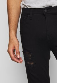 Svarta denimjeans med slitna detaljer på låret, med smal passform och standard femfackdesign. En hand vilar avslappnat i närheten.