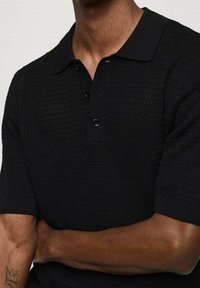 Homme portant un polo noir texturé à manches courtes avec un col et trois boutons, bras croisés, montrant un petit tatouage sur l'avant-bras gauche.