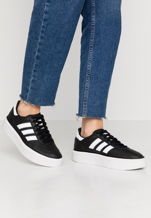 Person iført blå jeans og sorte sneakers med hvide striber og tykke hvide såler, stående på et lyst gulv.
