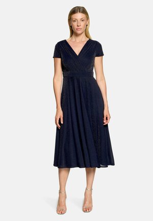 Frau trägt ein marineblaues, kurzärmeliges Midikleid mit V-Ausschnitt, tailliertem Schnitt und ausgestelltem Rock, steht vor einem schlichten Hintergrund.