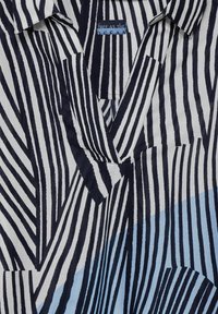 Maglietta con colletto a punte, caratterizzata da righe verticali blu navy e bianche con accenti blu chiaro e un tessuto texturizzato. L'etichetta reca la scritta "LIBERO COME L'OCEANO."