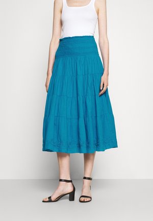 Femme portant un haut blanc sans manches, une jupe midi à volants bleu sarcelle avec taille smockée, et des sandales noires à talon carré avec bride à la cheville sur un fond clair.