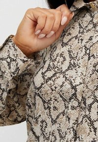 Ljusfärgad skjorta i ormskinnsmönster med en texturerad yta, med klassisk krage och knappdetalj, hålls av en hand med neutral nagellack.