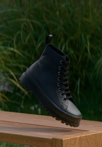 Botină din piele neagră la gleznă, cu vârf rotund, detalii cusute, talpă din cauciuc texturat și șireturi negre, poziționată deasupra unei suprafețe din lemn.