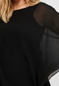 Haut noir avec des manches longues transparentes, un col rond et une silhouette fluide. Texture lisse avec un tissu léger et respirant.