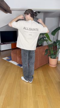 Gai bēšs pārdomāts T-krekls ar "ALLSAINTS" tekstu uz muguras, apvienots ar tumši pelēkiem džinsiem un baltām sporta apavām. Iekštelpu vide.