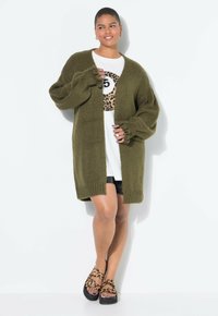 Olivgrüner, oversized Cardigan mit strukturiertem Strick und überschnittenen Ärmeln. Darunter ein weißes T-Shirt mit einem schwarz-braunen Leopardenmuster, kombiniert mit schwarzen Shorts und Schuhen im Tierdruck.