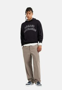 Felpa nera con la scritta "ARMANI EXCHANGE" in lettere bianche, abbinata a pantaloni beige e sneakers bianche con dettagli blu. Look e stile casual.