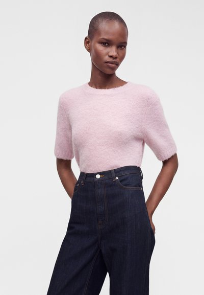 Mujer con un suéter corto de manga corta de color rosa claro y borroso y vaqueros azul oscuro de talle alto con las manos en los bolsillos sobre un fondo liso.