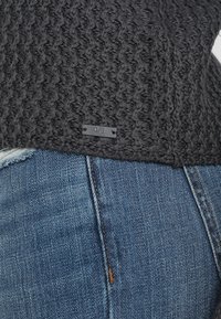 Chunky donkergrijze gebreide trui met een textuurpatroon en een klein zwart label aan de zoom, gecombineerd met blauwe denim jeans.