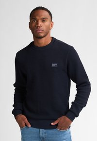 Marineblå sweatshirt i struktur med rund halsudskæring, ribbede manchetter og kant. Har et lille mærke på forsiden. Bæret sammen med lyseblå jeans.