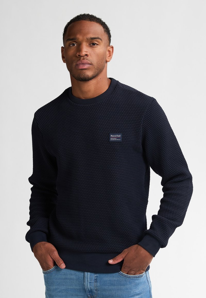 Marineblå sweatshirt i struktur med rund halsudskæring, ribbede manchetter og kant. Har et lille mærke på forsiden. Bæret sammen med lyseblå jeans.