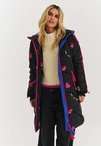 Černý prošívaný kabát s barevnými vyšívanými vzory, zapínáním na zip, modrými a růžovými doplňky a nastavitelnými šňůrkami v pase a na manžetách.