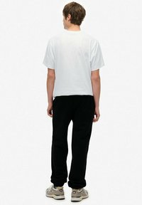 Weißes T-Shirt mit kurzen Ärmeln und schwarze Jogginghose, von hinten gesehen, mit Sneakers in Grau-Beige-Farbschema.