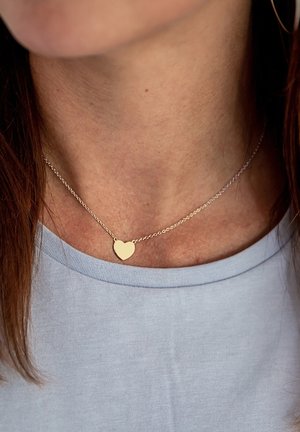 Goldenes Herz-Anhängerhalskette an einer feinen Kette, getragen auf einem hellblauen Hemd, das eine glatte Textur und ein einfaches Design zeigt.