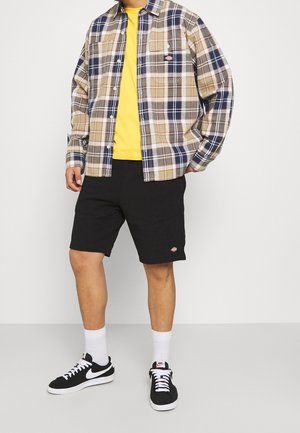 Man draagt een geruit beige en marineblauw overhemd over een geel T-shirt, zwarte korte broek, witte sokken en zwarte Nike sneakers, staande tegen een witte muur.