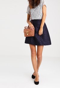 Weißes Oberteil mit Spitzenmuster, dunkelblaue knielange Rock mit strukturiertem Leopardenmuster, braune Handtasche mit Verschluss, schwarze flache Schuhe.