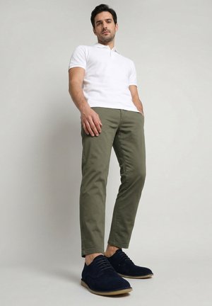 Muž v bílém polo tričku, olivově zelených kalhotách a tmavě modrých botách stojí před jednobarevným světle šedým pozadím.