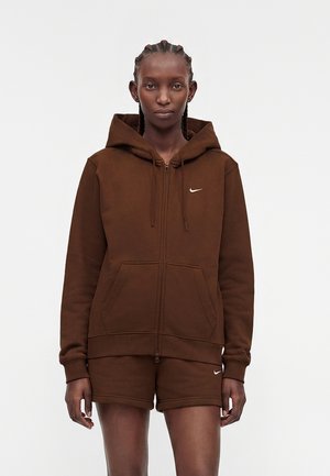 Kvinde med fletninger iført brun lynlås-hættetrøje og matchende shorts, begge med små hvide Nike swoosh-logoer.