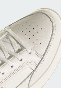 Zapatilla blanca de cuero con perforaciones, cordones planos y una suela de goma de color claro. Presenta costuras en contraste y una textura suave.