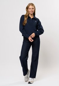 Mørkeblå, langermet zip-up hettegenser med ribbede mansjetter. Kombinert med mørke denimjeans og lys grå joggesko for et avslappet utseende.