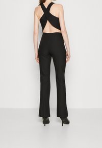 Svart jumpsuit med öppen rygg och korsade axelband; figurnära silhuett och utsvängda byxor. Bärs med svarta högklackade skor.