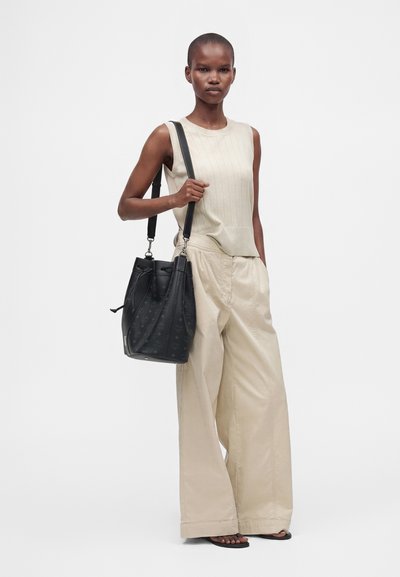 Femme portant un haut beige sans manches et un pantalon beige large, portant un grand sac à épaule en cuir noir, debout sur un fond uni.