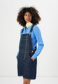 Robe salopette en denim, bleu foncé, avec des bretelles réglables et des poches avant. Associée à un haut à manches longues bleu clair. Texture lisse.