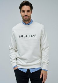 Sudadera de un beige claro con el logo "SALSA JEANS" en la parte frontal, puños acanalados y cuello redondo, superpuesta a una camisa de color azul claro con cuello.