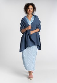 Niebieski szal o fakturze leżący na jasnoniebieskiej sukience w kropki. Modelka nosi różowe sandały na platformie, prezentując swobodny, warstwowy styl.