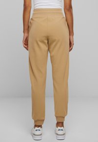 Beige joggebukser med tapered fit, elastisk linning og ribbede mansjetter. Stoffet ser mykt og glatt ut, brukt sammen med hvite treningssko.