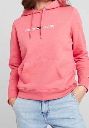 Kobieta w różowej bluzie z kapturem Tommy Jeans z kieszenią z przodu i sznurkami, w połączeniu z jasnoniebieskimi dżinsami, ręce w kieszeniach.