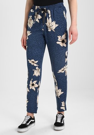 Niebieskie spodnie z czarnymi plamkami i dużym białym kwiatowym wzorem, elastyczna talia z czarnym sznurkiem, noszone z czarno-białymi trampkami.