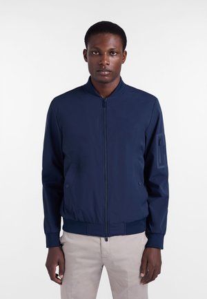 Uomo che indossa una giacca bomber blu navy con zip e tasche, e pantaloni beige, in piedi davanti a uno sfondo bianco semplice.