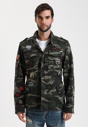 Giacca in cotone con motivo camouflage verde e nero, dotata di numerosi patch e distintivi, chiusura con bottoni e due tasche sul petto.