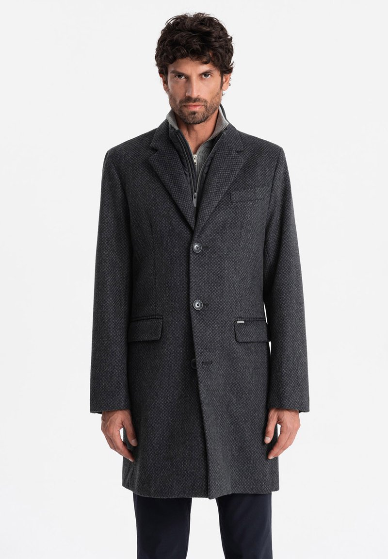 Abrigo gris de mezcla de lana con un patrón texturizado, solapas tipo notch, dos bolsillos frontales y tres botones. Llevado sobre una chaqueta gris con cremallera.