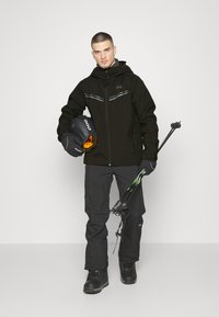 Schwarze Skijacke mit reflektierenden Akzenten, kombiniert mit schwarzen Skihosen und Handschuhen. Hält einen schwarzen Helm und Skistöcke mit grünen Akzenten.