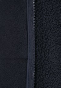 Gros plan sur un vêtement en polaire bleu marine foncé montrant un tissu texturé et doux à droite et un tissu plus lisse à gauche, séparés par une fermeture éclair en plastique.