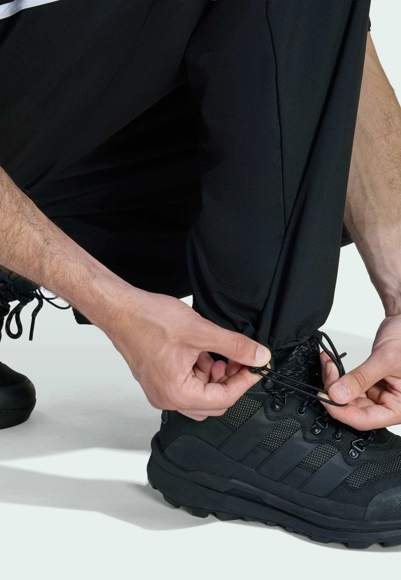 Zwarte wandelschoen met een gestructureerde bovenkant en rubberen zool. Handen die de veters verstellen, terwijl ze zwarte broek dragen met een lichte elastische cuff bij de enkel.