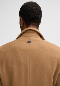 Bruine wollen overjas met een brede kraag, voorzien van een achterlabeldetail. De stof heeft een zachte textuur, wat zorgt voor een nette, op maat gemaakte uitstraling.