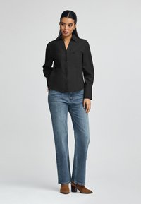 Svart skjortblus med krage, bröstficka och långa ärmar, kombinerad med ljusblå raka jeans och bruna klackstövlar.