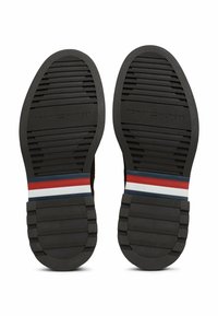 Musta kumipohja, jossa on urittettu pinta. Värikorostuksia punaisena, valkoisena ja sinisenä kantapään lähellä. "TOMMY HILFIGER" on painettu pohjaan.